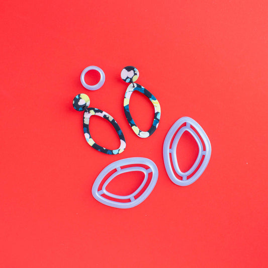 2 mirrored oval shaped cutters and a stud together with a set of finished polymer clay earrings on a red background.