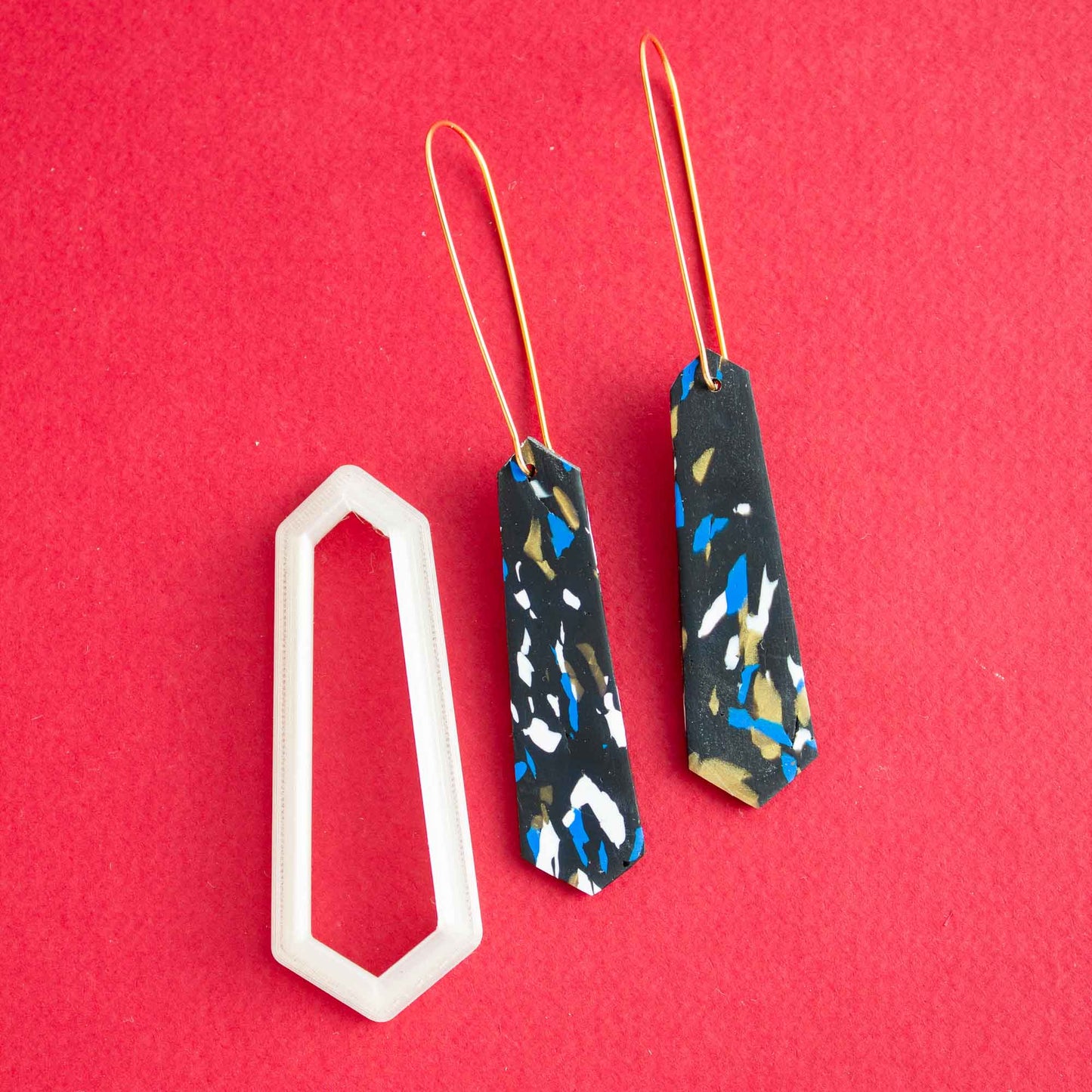 1 pair of long thin earrings next to a polymer clay cutter of the same shape on a red background