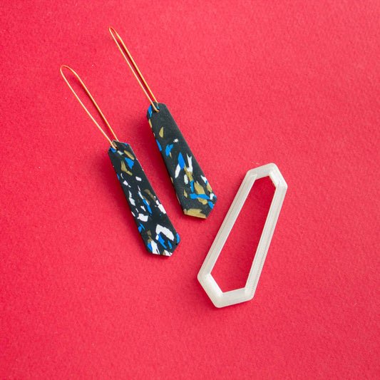 a pair of long thin earrings next to a polymer clay cutter of the same shape on a red background