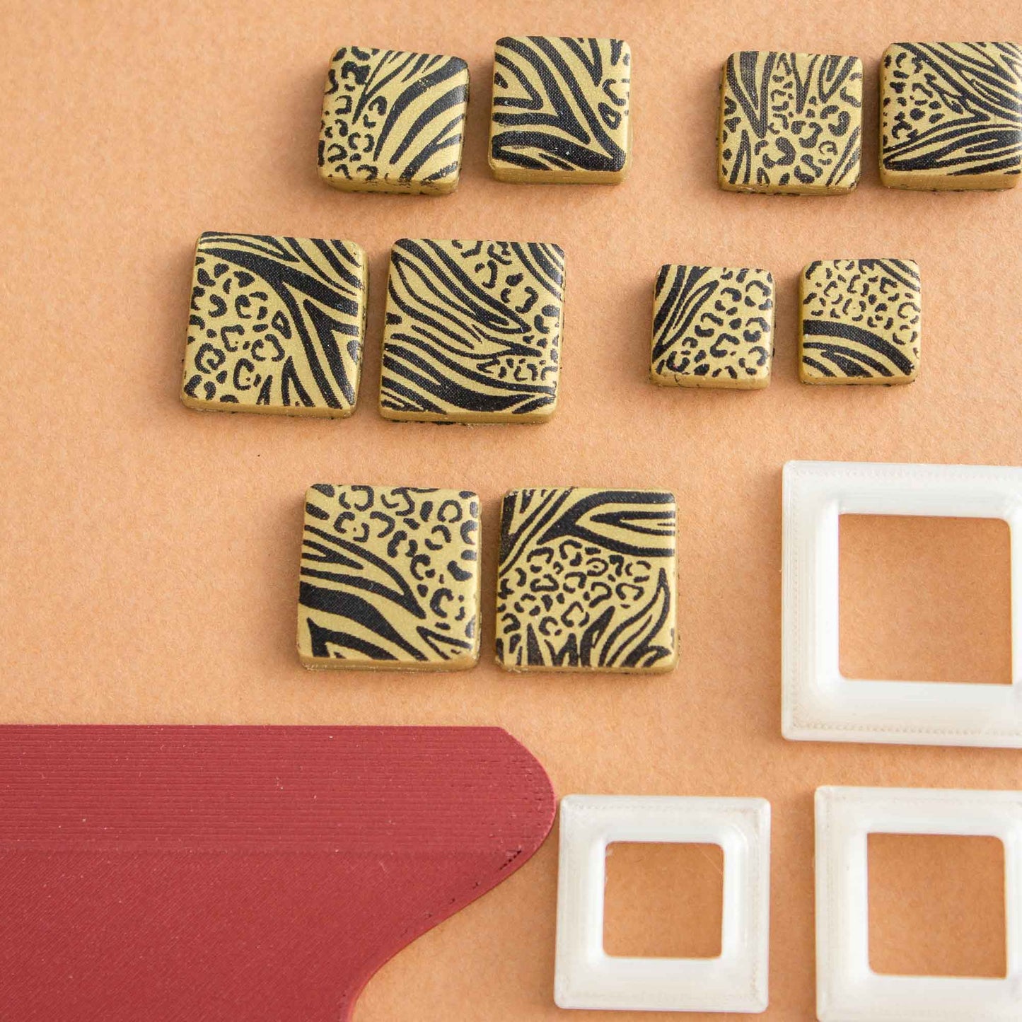 close up of 1 Animal pattern small silkscreen, some square animal printed polymer clay shapes, 3 square cutters for clay and one squeegee on a light brown background