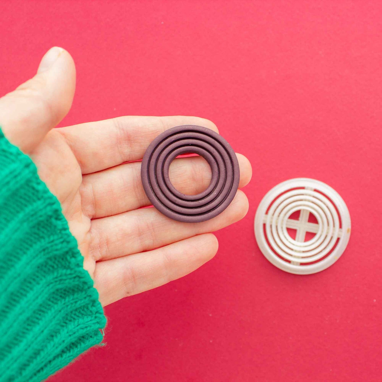 Circle shape with inside circle patterns on a hand in a red background with a polymer clay cutter of the same shape